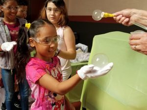 Young girl wearing safety goggles participating in a science project including bubbles as part of a summer learning program