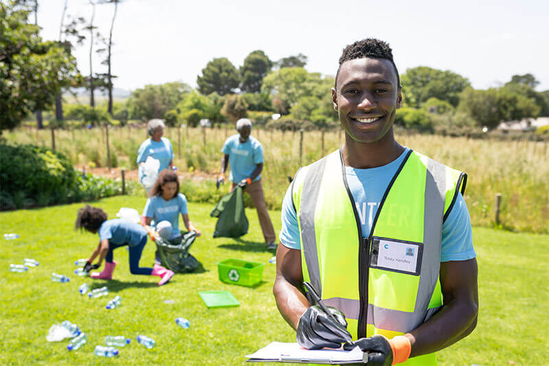 Volunteer smiling directly at camera wearing a yellow safety vest and clip board with volunteers cleaning up trash behind him