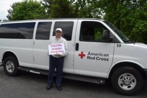 An older man is standing in front of a Red Cross van holding a sign that says United Way Changes Lives
