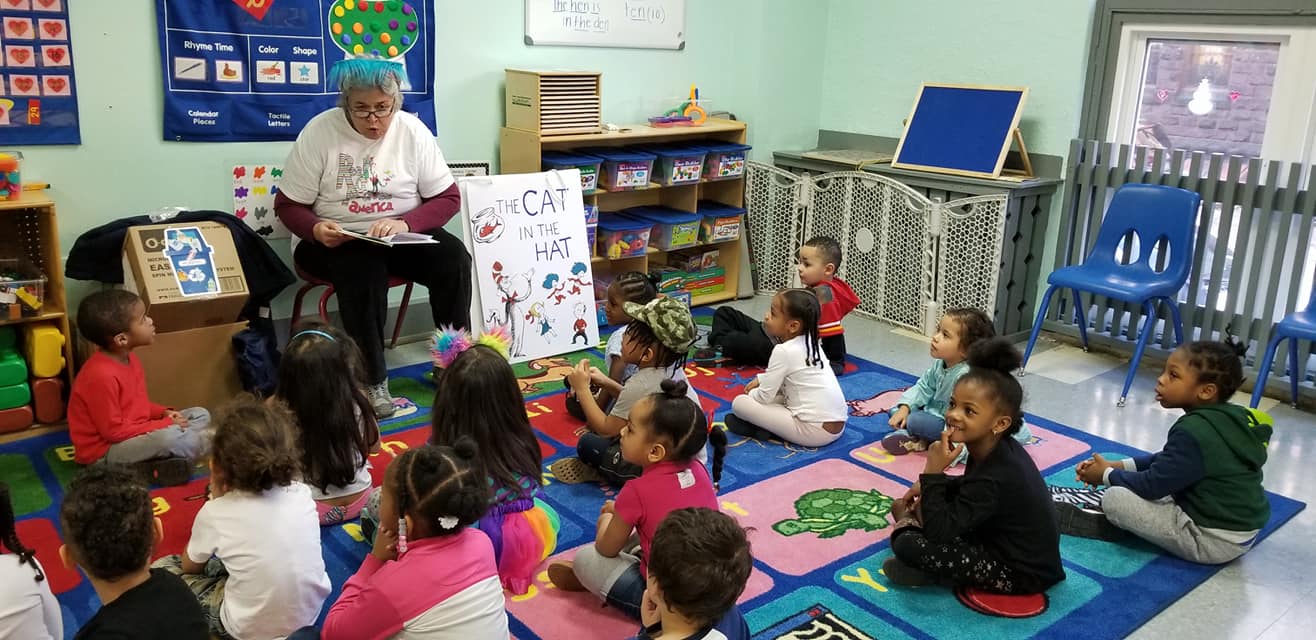 Read Across America Day volunteer in blue wig reading The Cat in the Hat to children in a classroom