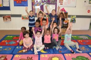 a group of young children in a preschool classroom smiling and waving their hands in the air