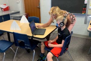A boy is sitting at a table in a classroom. He is wearing headphones and looking at a laptop. A teacher is bending near the boy and pointing at the laptop
