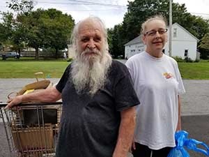 A man with a long beard and a woman are smiling at the camera. The man has his hand on a cart full of groceries from a food pantry