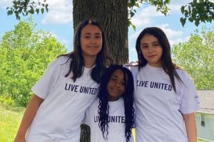 Three girls are standing in front of a tree. The girls are wearing Live United t-shirts and have their arms around each other