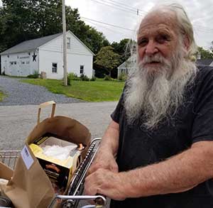 An 84-year-old man with a long white beard is standing next to a grocery cart full of fresh groceries obtained at a food pantry in Kutztown, PA