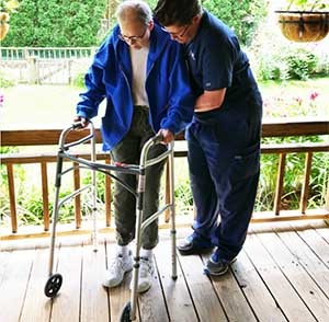 A woman is using a walker on her deck while a physical therapist helps her