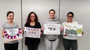 Four women standing in a line indoors. Each woman is holding a handmade "thank you" sign