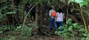 Two people with their backs to the camera are walking into the woods. One is a carrying an orange backpack. They are medical professionals walking into the woods to provide medical care to homeless people.