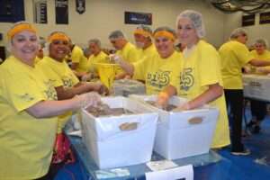 A group of women wearing bright yellow t-shirts and hair nets are smiling at the camera as they package meals