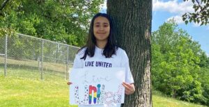 a teenage girl is standing by a tree. she is holding a sign that says "Live United"