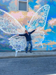 A man is standing in front of a butterfly mural painted on the side of a building. He is standing between the wings of the butterfly