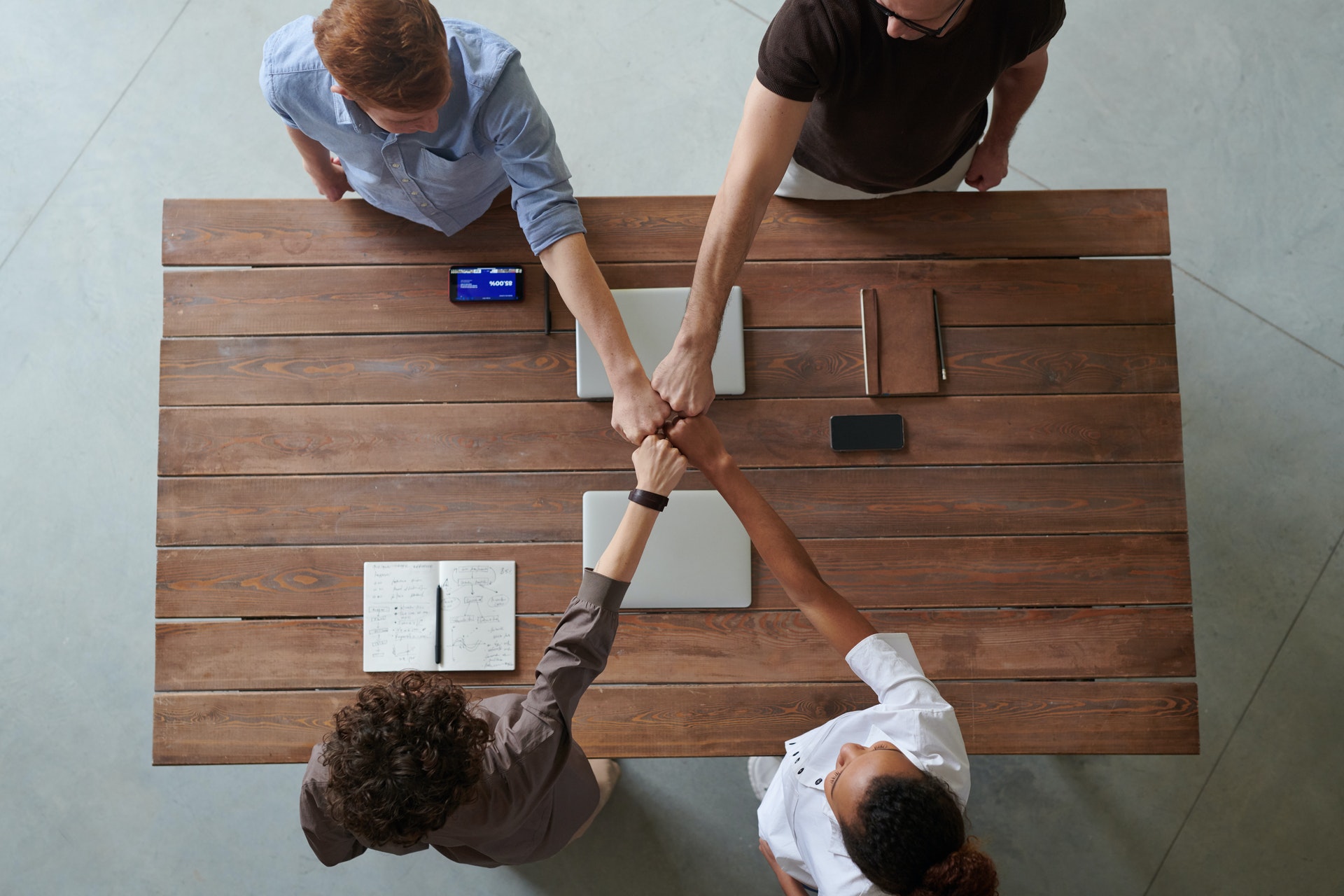 Four adults are standing around a wooden table. They are doing a four-person fist bump in the center of the table. Photo by fauxels: https://www.pexels.com/photo/photo-of-people-doing-fist-bump-3184430/