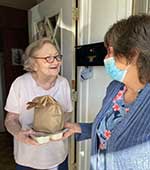 An older women is standing in the doorway of her home. She is accpeting a brown bag and a small tray of food from another woman