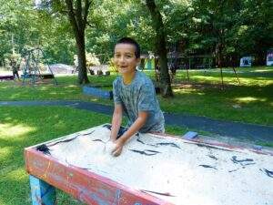 A boy of about 10 or 11 years old is smiling at the camera. He is standing outside in a wooded area and his hands are in a sand sensory box