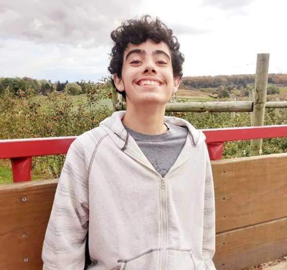 A young teenage boy with short culry black hair is smiling a the camera. He is wearing a zip-up hoodie and a grey t-shirt. He is sittingin the back of a hay wagon