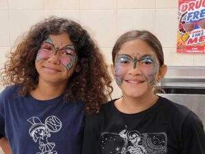 Two girls are standing side-by-side in front of a tiled wall. Each girl has a butterfly painted on her face. Both girls are smiling