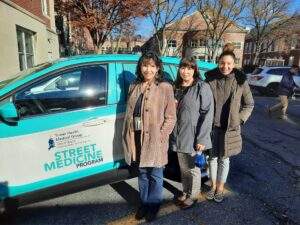 Three women wearing winter coats are standing outside in front of a white and teal SUV with the Street Medicine logo on the door