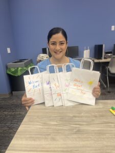 A woman is smiling and holding up a variety of gift bags with inspirational messages hand-written in marker