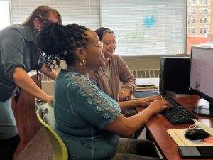 A woman is sitting in profile at a computer and smiling. A man is standing behind her and another woman is sitting beside her