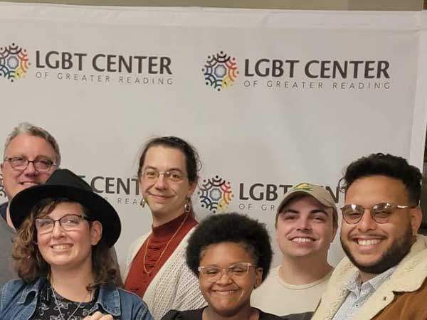 A group of people of various ages are standing in front of a backdrop with the LGBT Center logo on it