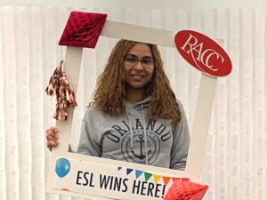 A young woman is holding a large Instagram-type photo frame with the RACC logo and the words ESL Lives Here