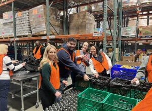 Four people wearing orange safety vests are packing food into black bags in a food bank warehouse