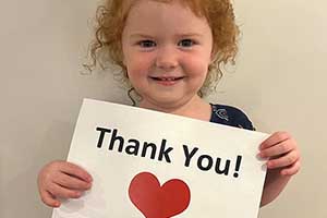 A pre-school-aged girl is smiling at the camera and holding a thank you sign