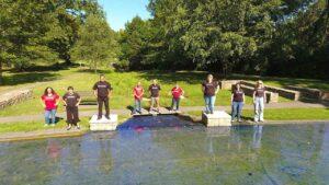 A group of nine men and women are standing with hands on hips, facing a pond and smiling