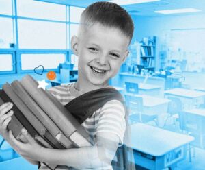 A boy is standing in a classroom. He is holding a stack of books and smiling