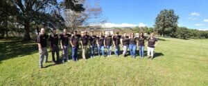 A diverse group of men and women are standing in a grassy field. They are wearing ct-shirts that say "It Starts with U". Each person has placed their elbow on the shoulder of the person next to them