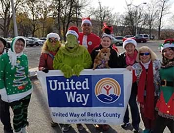 A group of men and women wearing Santa hats are standing behind a large United Way banner. The Grinch is also standing with the group