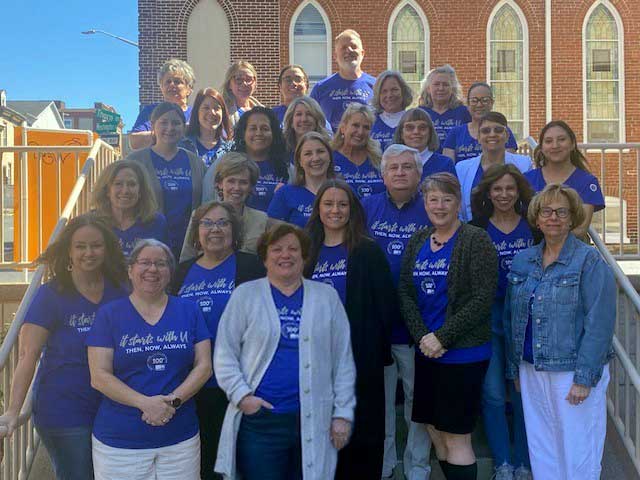 staff on steps w Jean for web United Way staff standing outside and wearing blue shirts that say "it starts with U" and feature the 100th anniversary logo