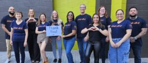 A group of men and women wearing dark blue t-shirts that say "it starts with u" are standing outside near an old brick building. Two women in the center are holding a sign that says "thank you"