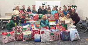 A group of women are gathered together in a conference room, surrounded by piles gifts in brightly colored Christmas bags