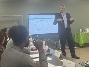A tall man is presenting at the front of a conference room. A man is sitting in the foreground of the photo and listening