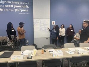 Three women are standing a large conference room. One of the women is pointing to a large piece of paper on the wall. A man in a hat is looking at what she is pointing at