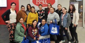A group of women are gathered together in front of a sign that says RACC. Some of them are holding blue bags with the United Way of Berks County logo. All are smiling