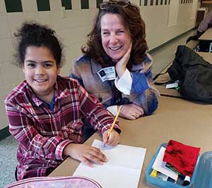 A young girl sits with her tutor in a classroom. They're both smiling