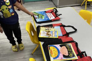A child's hand is reaching to a book on a table. There are three red bags lying side by side on the table. Each bag has a children's book lying on top of it