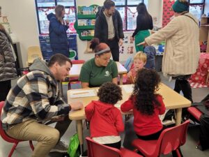 A group of adults are in a child care classroom. One man is seated and talking with two children, who are facing away from the camera