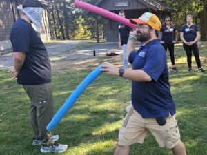 Two men are outside on a sunny Fall day. They are participating in a teambuilding activity. One man has two poodles. He is approaching a man wearing a blindfold.