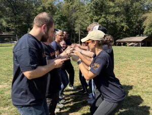 A group of men and women are standing outside in two lines. They are facing each other and holding a board with their fingertips in a team building exercise