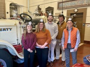 A group of men and women are standing in front of an old-fashioned fire truck