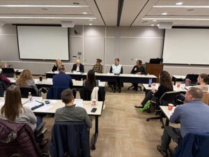 A group of men and women are sitting a long table at the front of a classroom-type room. They are participating in a panel discussion