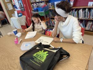 An adult volunteer and a young student work together at a wooden table as part of the Ready.Set.READ! program. A black tote bag with the program's logo sits in the foreground, while the background shows colorful library bookshelves. The student is focused on a worksheet while the tutor observes, with a small whiteboard nearby showing the words "music notes."