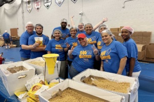 A diverse group of volunteers wearing blue "Big Cheese" t-shirts, hairnets, and gloves pose for a photo behind tables filled with large boxes of dry macaroni. The setting is a gymnasium where the volunteers are participating in a United Way meal-packaging event.