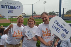 Three volunteers wearing white "Day of Caring" t-shirts smile while holding United Way signs at a baseball stadium. The signs feature slogans such as "I MAKE A DIFFERENCE" and "I AM CHANGING LIVES," with the United Way of Berks County logo visible against a background of a baseball field and stadium seating.