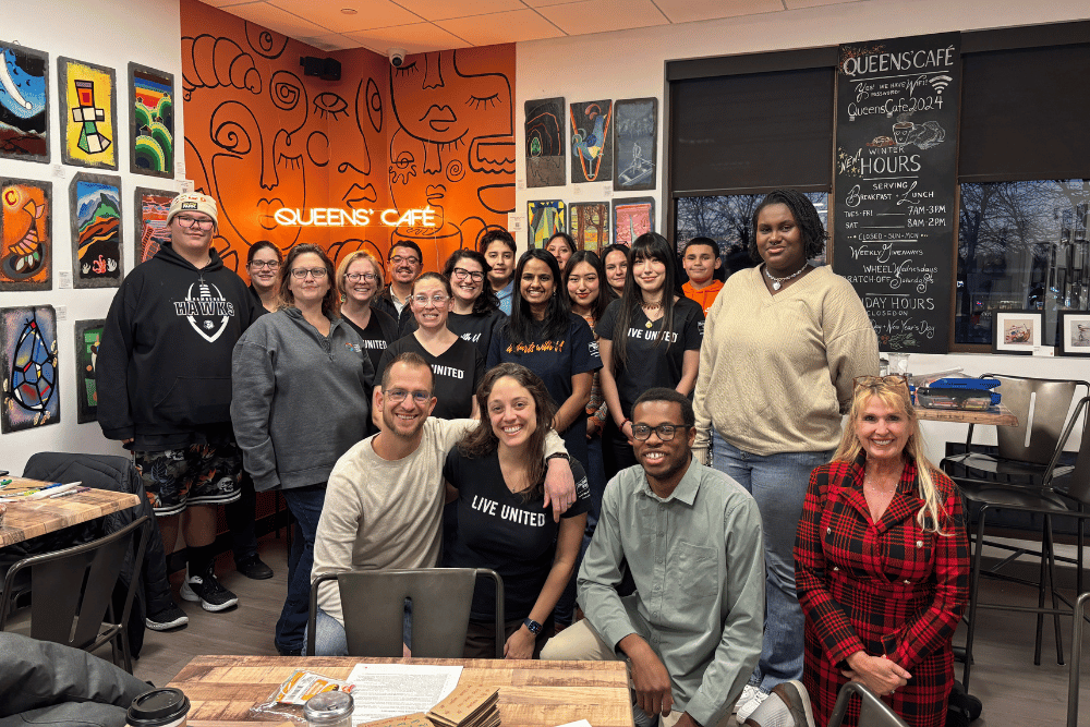 A group shot of members from the United Way of Berks County Emerging Leaders program posing together inside Queen's Cafe. The group is arranged in front of a bright orange mural and a wall displaying various framed artworks. Several individuals are wearing black t-shirts that say "LIVE UNITED." In the background, a neon "Queen's Cafe" sign and a chalkboard menu are visible.
