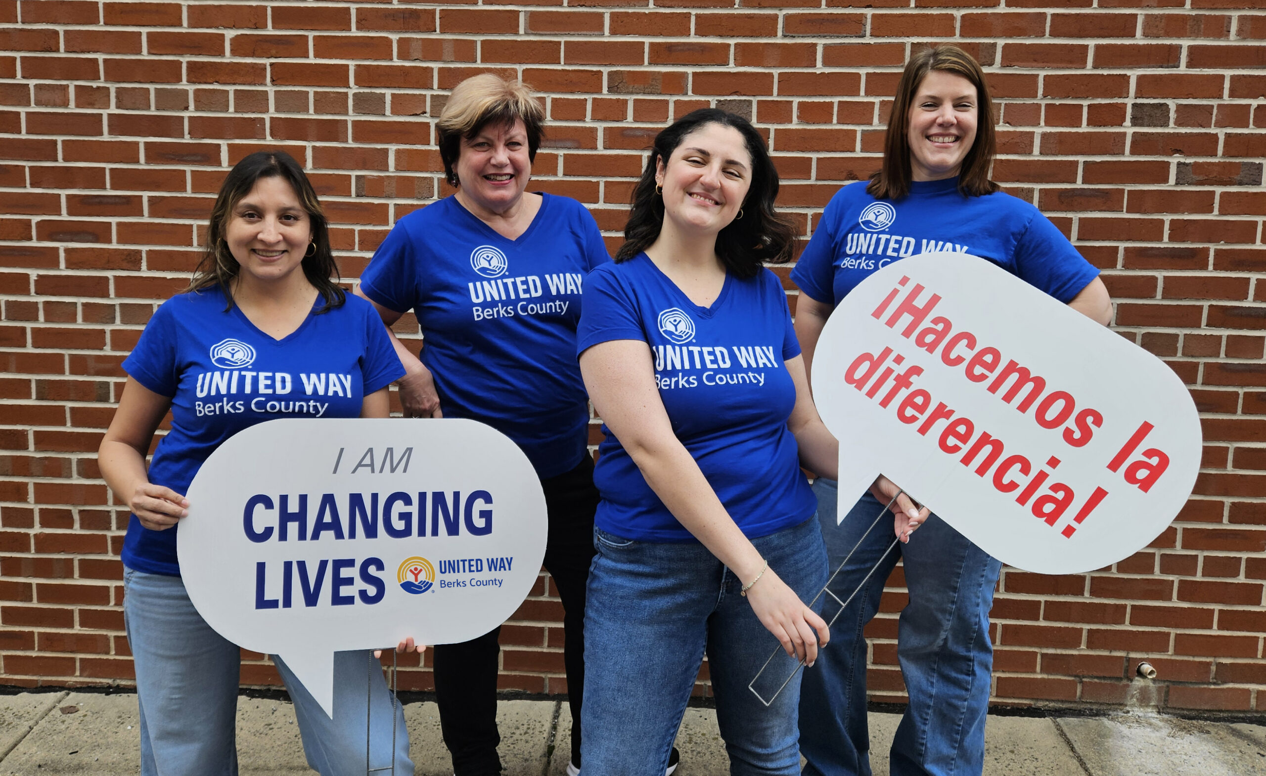 Gemini said Three volunteers wearing white "Day of Caring" t-shirts smile while holding United Way signs at a baseball stadium. The signs feature slogans such as "I MAKE A DIFFERENCE" and "I AM CHANGING LIVES," with the United Way of Berks County logo visible against a background of a baseball field and stadium seating.