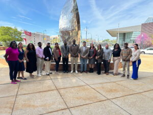 A group of fifteen people from the United Way of Berks County Leadership United program stands in a line for a group photo in a sunny outdoor plaza. Behind them is a large, reflective, flame-shaped metallic sculpture. In the background, city buildings—including one with a 'RACC' sign—and a clear blue sky are visible.